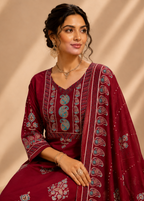 A woman wearing a deep maroon traditional Indian outfit with intricate paisley and floral embroidery in light blue, white, and gold, posed against a warm beige background with soft diagonal shadows.