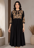 A woman modeling a black midi dress with gold floral embroidery on the bodice and three-quarter sleeves, standing in a neutral indoor setting.