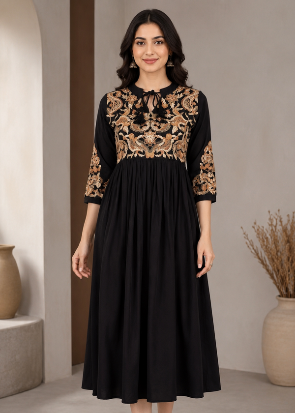 A woman modeling a black midi dress with gold floral embroidery on the bodice and three-quarter sleeves, standing in a neutral indoor setting.