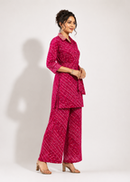 A woman modeling a bright pink two-piece kurta set with a white bandhani pattern, consisting of a long-sleeve collared top and matching wide-leg pants, set against a plain light gray studio background.