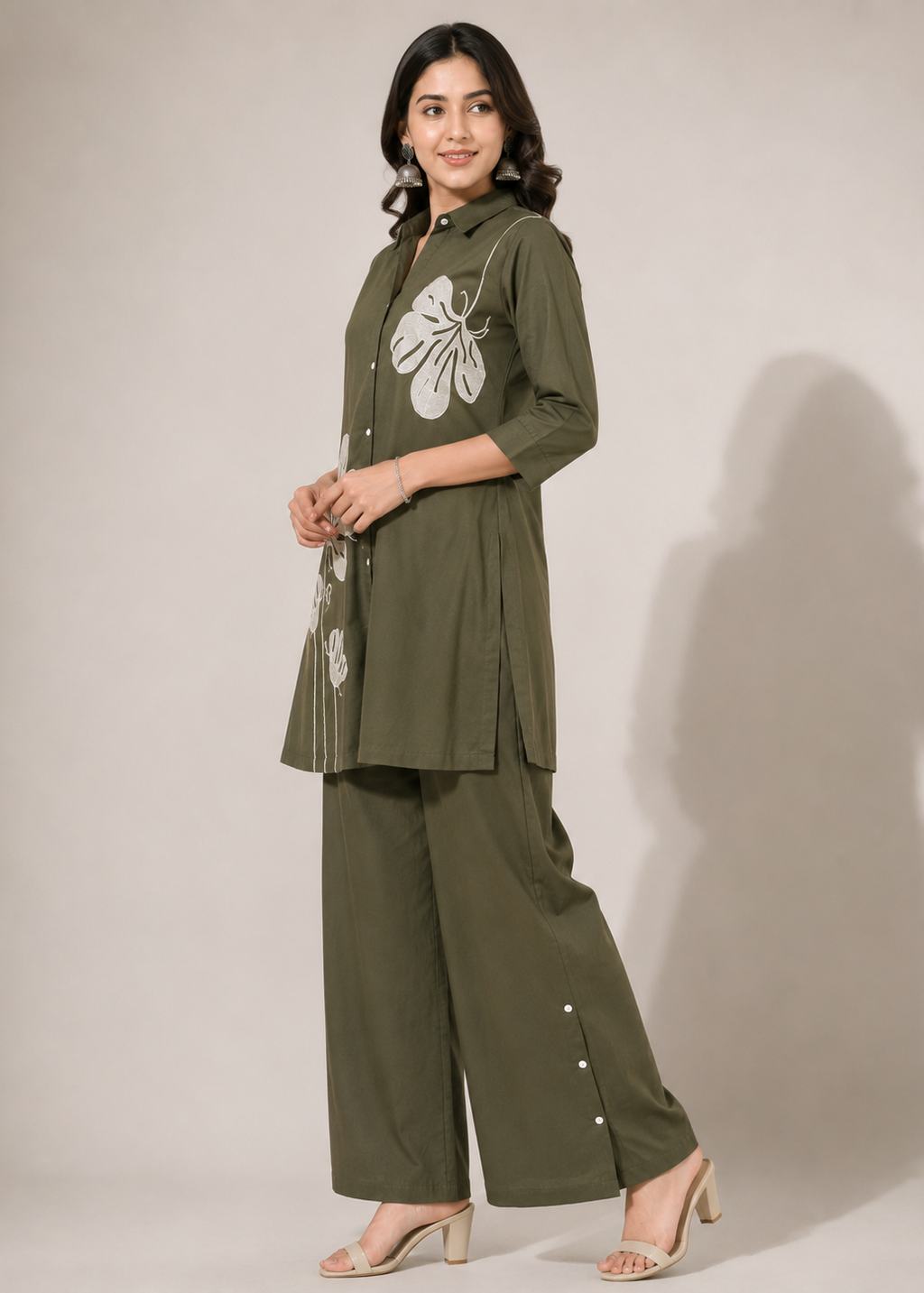 A woman modeling an olive green two-piece kurta and trouser set with white floral embroidery on the chest and side, paired with beige heeled sandals against a plain studio background.