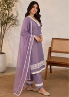 A woman models a lavender purple ethnic suit set with white lace trim, consisting of a kurta, pants, and a dupatta, in a styled indoor setting with a wooden chair and potted plant.