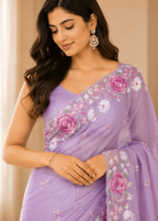 A woman wearing a lavender purple saree with pink and white floral embroidery, paired with a matching embroidered blouse, set against a soft beige background.