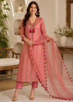 A woman models a pink traditional Indian salwar kameez set with a matching sheer dupatta. The set features a pink embroidered kurta with a deep V-neck, pink trousers, and a matching pink dupatta adorned with red pom-poms along the border.