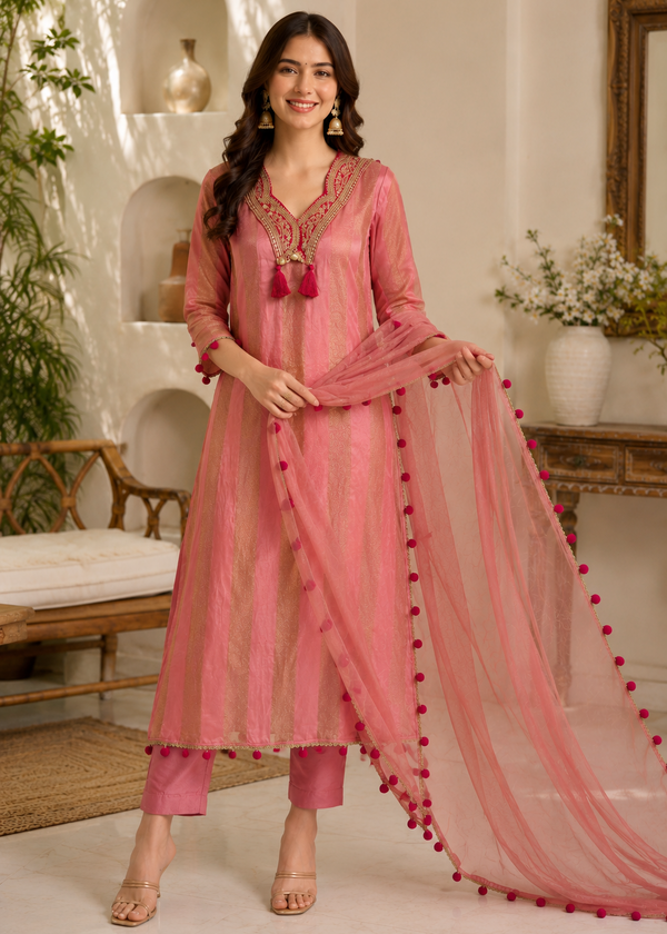 A woman models a pink traditional Indian salwar kameez set with a matching sheer dupatta. The set features a pink embroidered kurta with a deep V-neck, pink trousers, and a matching pink dupatta adorned with red pom-poms along the border.