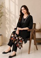 Woman sitting on a chair wearing a black outfit with a patterned scarf in a bright room.