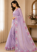 A woman models a lavender purple saree with pink and white floral embroidery along the border and pallu, set against a warm beige indoor background with curtains and a console table.
