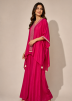 A woman modeling a bright magenta pink traditional outfit consisting of a long kurta with silver embroidery and a matching draped shawl with tassel details, set against a plain beige studio background.