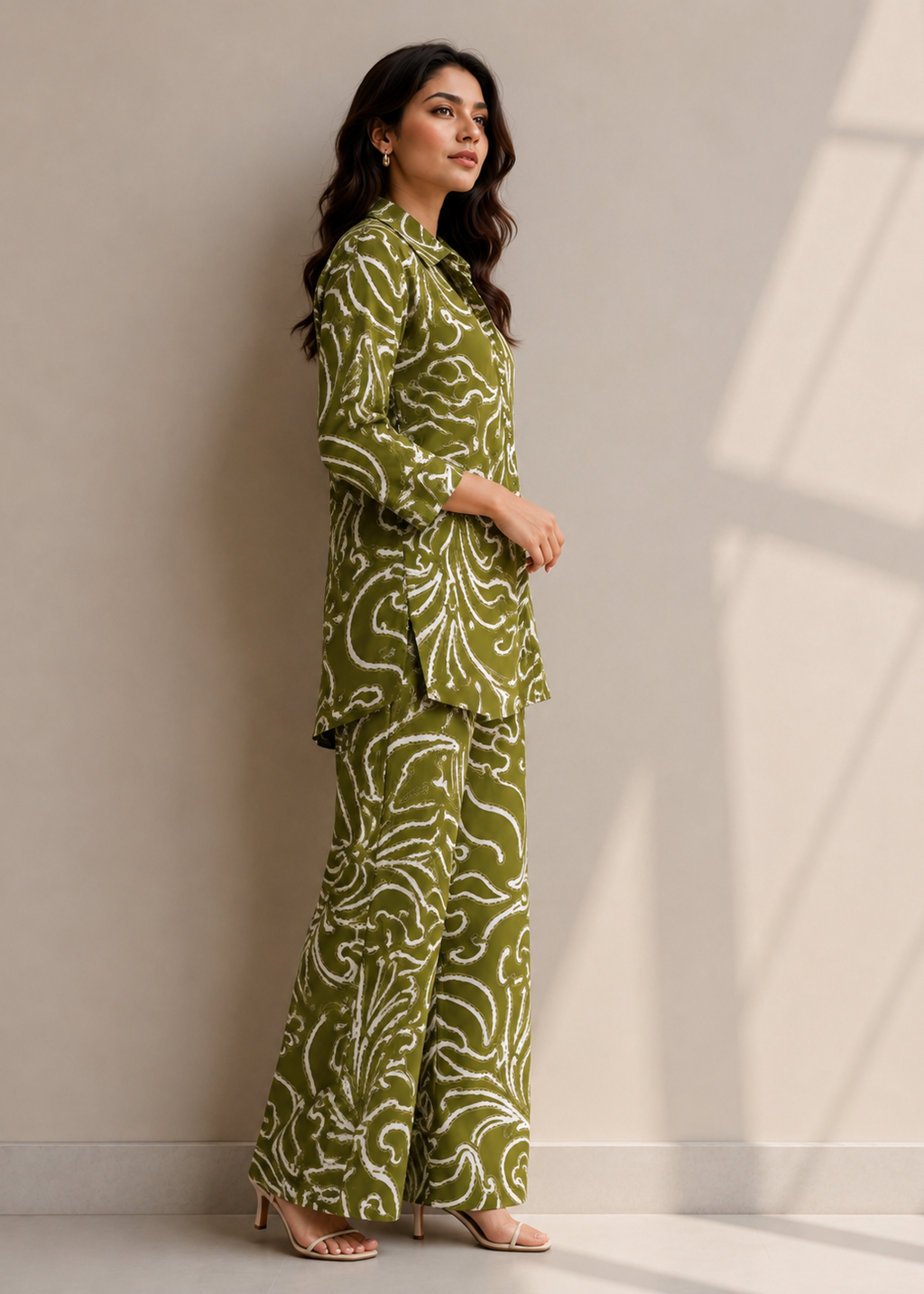 A woman modeling a matching olive green and white patterned two-piece outfit consisting of a long-sleeve collared top and wide-leg trousers, standing against a neutral beige wall with soft window shadow patterns.