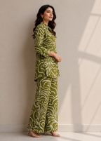 A woman modeling a matching olive green and white patterned two-piece outfit consisting of a long-sleeve collared top and wide-leg trousers, standing against a neutral beige wall with soft window shadow patterns.