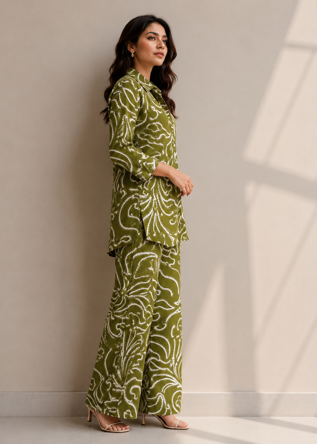 A woman modeling a matching olive green and white patterned two-piece outfit consisting of a long-sleeve collared top and wide-leg trousers, standing against a neutral beige wall with soft window shadow patterns.