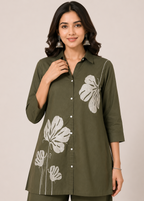 A woman wearing an olive green button-up tunic top with white floral prints, paired with matching olive green pants, set against a plain beige studio background.