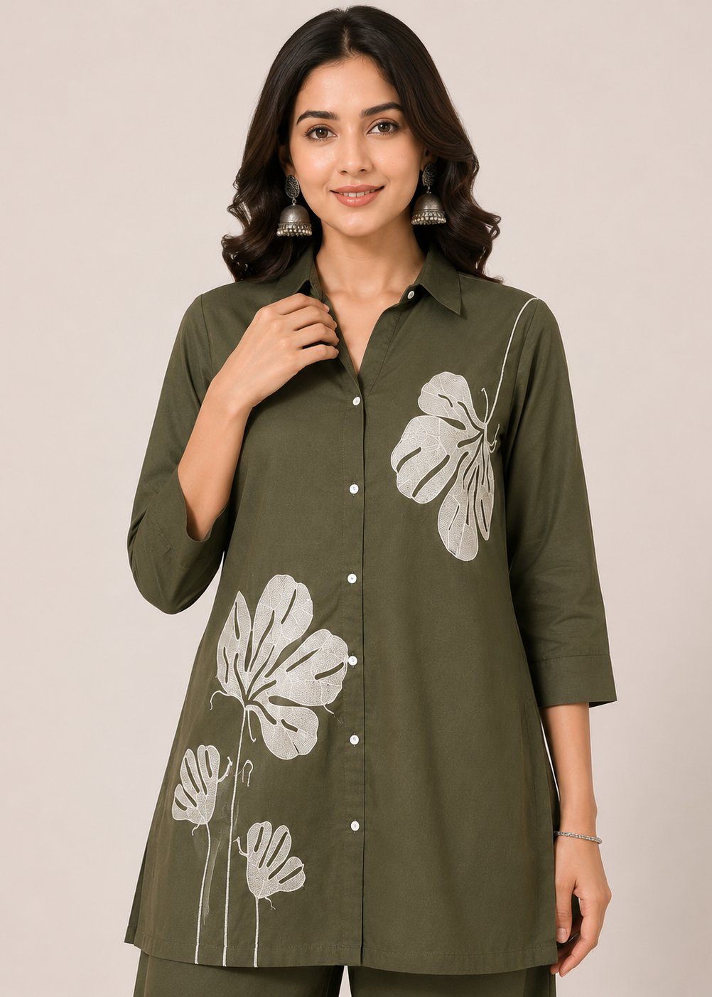 A woman wearing an olive green button-up tunic top with white floral prints, paired with matching olive green pants, set against a plain beige studio background.