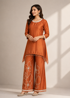 A woman modeling a rust-orange kurta and wide-leg pants set with white floral embroidery and tassel trim, standing against a neutral beige wall with soft window shadow patterns.