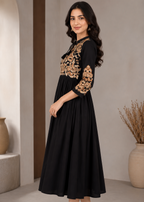 A woman modeling a black midi dress with gold floral embroidery on the bodice and three-quarter sleeves, paired with beige heeled sandals in a neutral indoor setting.
