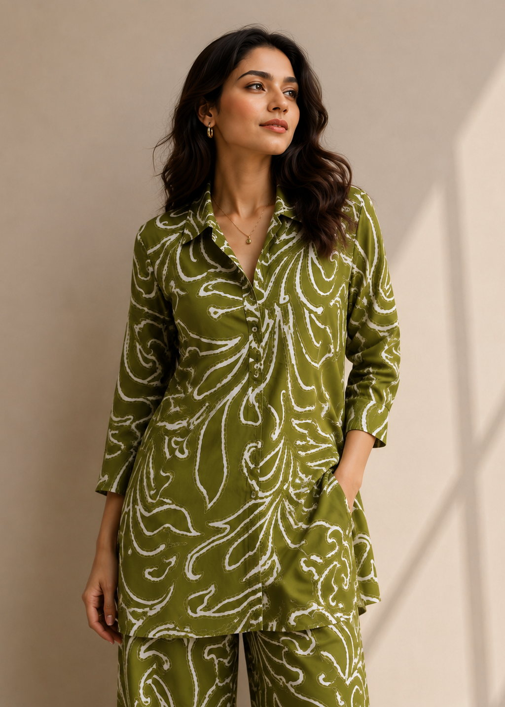 A woman modeling a matching olive green two-piece pajama set with a white abstract floral pattern, standing against a neutral beige wall with soft shadows.