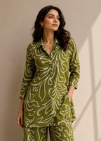 A woman modeling a matching olive green two-piece pajama set with a white abstract floral pattern, standing against a neutral beige wall with soft shadows.