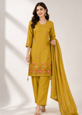 A woman modeling a mustard yellow ethnic three-piece suit with pink floral embroidery on the kurta and matching dupatta, set against a neutral studio background with soft window shadows.
