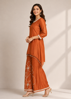 A woman modeling a rust-orange traditional Indian outfit with matching wide-leg pants, featuring white floral embroidery and gold accents, set against a neutral studio background with soft window shadow patterns.
