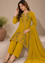 A woman models a mustard yellow ethnic three-piece suit consisting of a long kurta with green and gold embroidery, matching wide-leg trousers, and a draped dupatta, set against a neutral beige background.