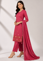 A woman models a three-piece salwar kameez suit in a vibrant magenta pink, featuring a long-sleeved embroidered kameez top, matching cropped trousers, and a sheer dupatta with gold and pink floral embroidery.