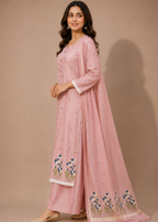 A woman modeling a light pink traditional outfit with matching dupatta, featuring blue and green floral embroidery near the hem and white contrast trim on the sleeves and border, set against a solid beige studio background.