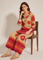 A woman sits on a wooden chair wearing a beige two-piece set with red and orange circular and geometric patterns, consisting of a long-sleeve button-up shirt and matching wide-leg pants.