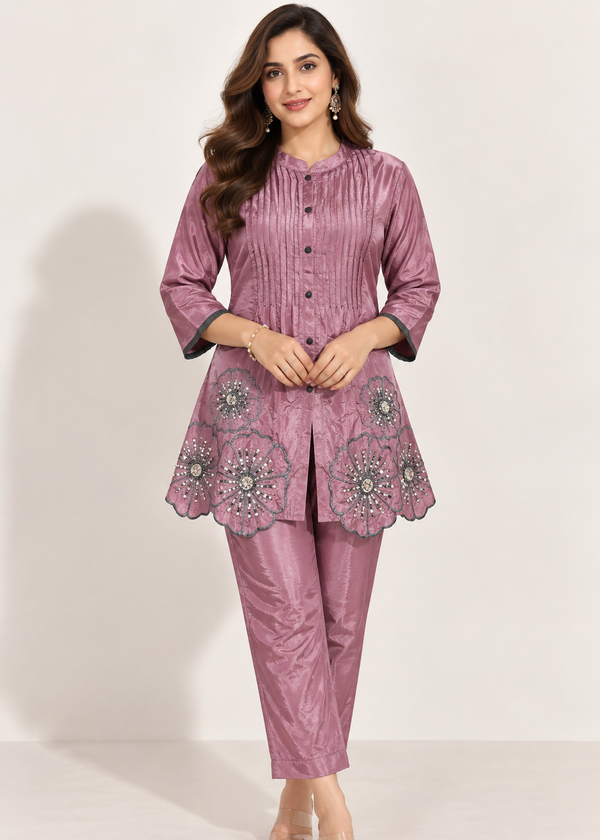 A woman modeling a two-piece salwar kameez set in dusty rose pink. The set features a long-sleeved tunic with a pleated front, button details, and a flared hem decorated with intricate floral embroidery, paired with matching slim-fit trousers.
