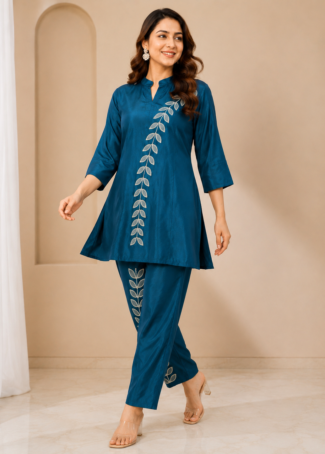 A woman modeling a teal blue two-piece kurta set with white leaf embroidery, standing in a beige indoor setting.