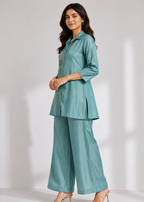A woman modeling a matching teal two-piece outfit consisting of a long-sleeve button-up kurta and wide-leg trousers, styled with nude high-heeled sandals against a light gray studio background.