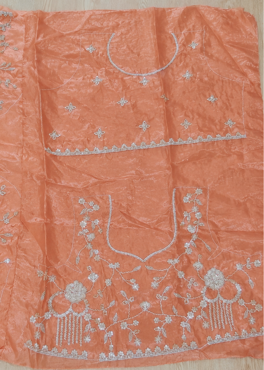An orange peach-colored fabric garment laid flat on a light wood surface, featuring intricate silver and white embroidery with floral motifs and mirror work.