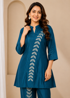 A woman modeling a teal blue kurta with a vertical white leaf embroidery design down the front, paired with matching trousers, set against a warm beige background with an arched alcove.