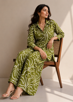 A woman sits on a wooden chair wearing a green and white patterned long-sleeve shirt and matching wide-leg pants. The setting is a minimalist studio with warm beige walls and floor, with soft shadows cast across the scene.