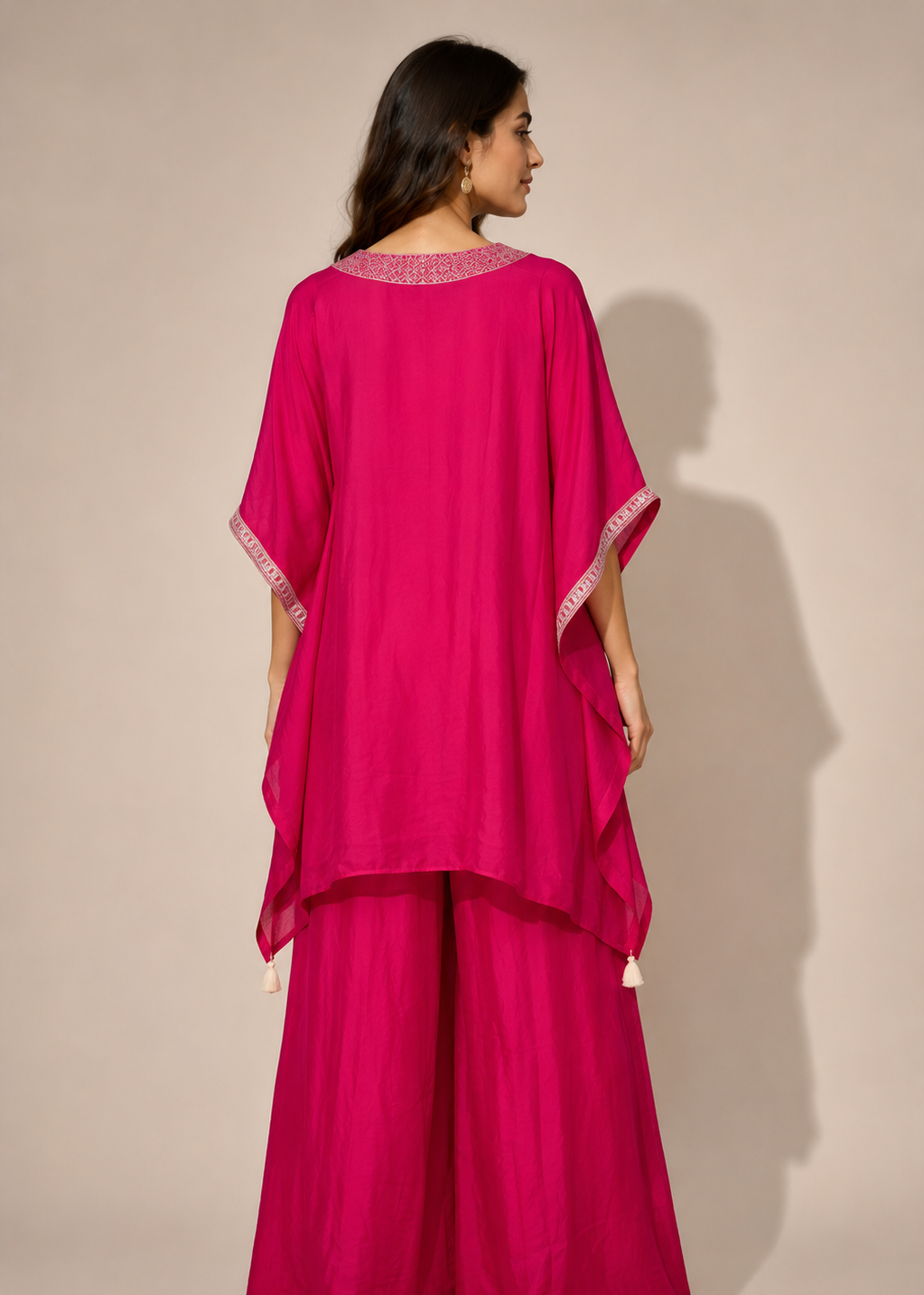 A woman shown from the back wearing a bright fuchsia magenta traditional outfit with wide draped sleeves and matching wide-leg pants, photographed against a plain beige studio background.