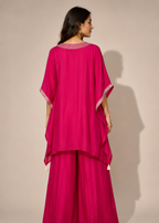 A woman shown from the back wearing a bright fuchsia magenta traditional outfit with wide draped sleeves and matching wide-leg pants, photographed against a plain beige studio background.