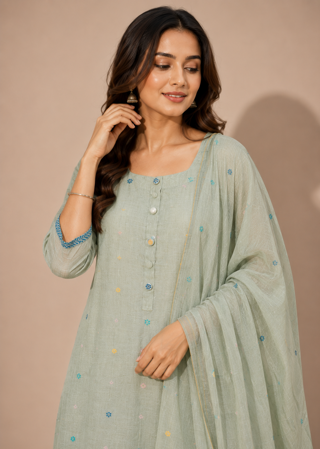 A woman wearing a light sage green traditional outfit with small blue and yellow embroidered details, paired with a matching sheer dupatta, set against a solid beige background.