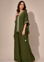 A woman modeling a dark olive green two-piece outfit consisting of a loose, draped tunic top with light green patterned trim and wide-leg pants, set against a solid beige studio background.