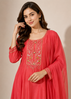 A woman modeling a coral pink traditional Indian kurta with gold and green floral embroidery on the chest, paired with a matching sheer dupatta, set against a solid beige studio background.