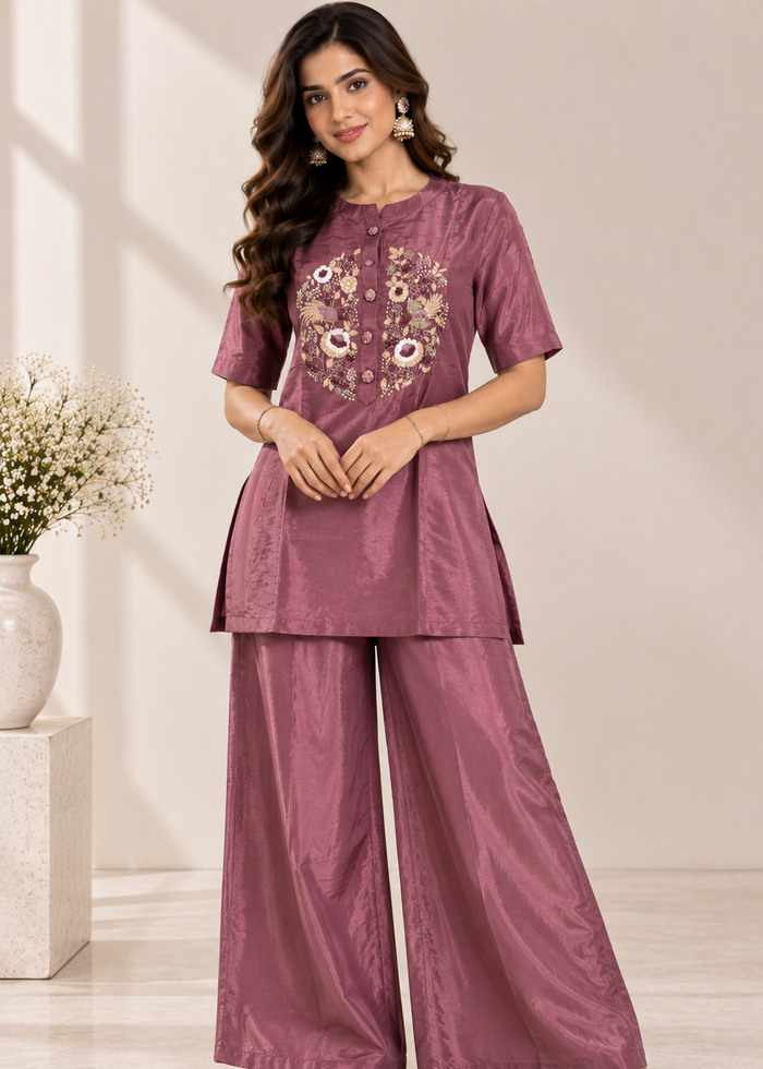 A woman models a two-piece salwar kameez set in a dusty rose/mauve color. The set features a short-sleeve tunic top with floral embroidery on the chest and matching wide-leg trousers.
