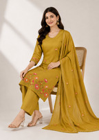 A woman models a mustard yellow ethnic salwar kameez suit with floral embroidery on the kurta and matching dupatta, seated on a wooden chair against a neutral studio background.