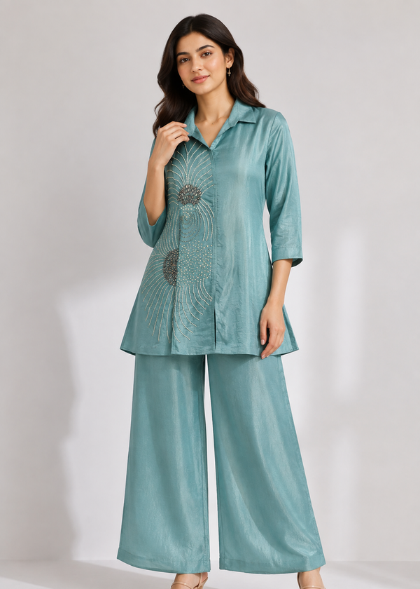 A woman modeling a matching two-piece outfit consisting of a light teal-green satin kurta with a floral mandala print on the front and wide-leg trousers, set against a plain light gray studio background.
