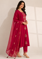A woman modeling a deep magenta red traditional Indian outfit, featuring a long kurta with gold embroidery, matching pants, and a sheer dupatta with paisley motifs, set against a plain off-white background.