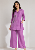 A woman modeling a matching purple two-piece outfit consisting of a long-sleeve collared tunic top with embroidered detailing and wide-leg trousers, photographed against a plain light gray studio background.