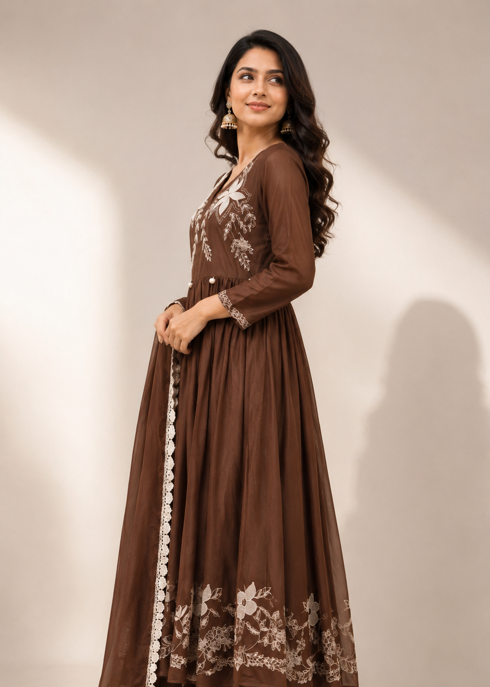 A woman modeling a long brown anarkali-style dress with beige floral embroidery and white border trim, paired with matching pants and heels, against a plain beige studio background.