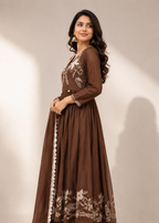 A woman modeling a long brown anarkali-style dress with beige floral embroidery and white border trim, paired with matching pants and heels, against a plain beige studio background.