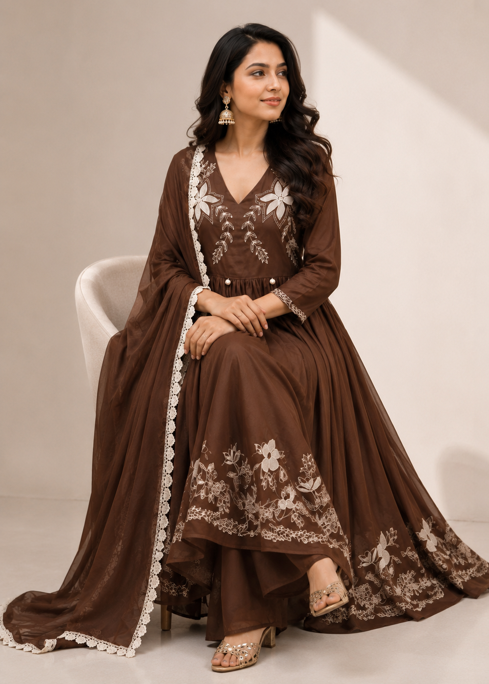 A woman seated on a white chair wearing a brown traditional Anarkali-style dress with white floral embroidery and a matching draped scarf, photographed in a clean studio setting.