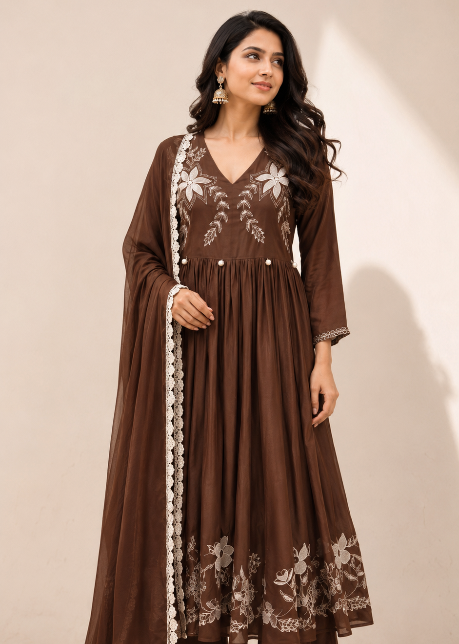 A woman models a brown traditional Anarkali kurta set with white floral embroidery and a matching dupatta draped over her shoulder, set against a plain beige studio background.