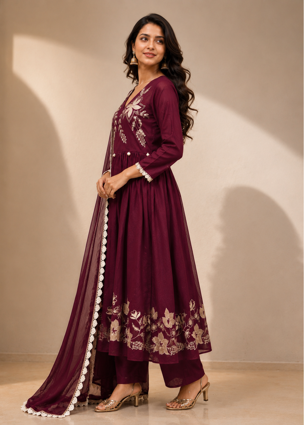 A woman modeling a deep maroon anarkali suit with beige floral embroidery and white border details, paired with matching pants and a sheer dupatta, set against a warm beige wall with soft shadows.