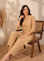 A woman sits on a wooden chair wearing a beige two-piece kurta set with a matching top and trousers, featuring a subtle floral print on the front. The setting is a warm, neutral-toned room with a woven rug, a pampas grass vase, and soft leaf shadows on the wall.