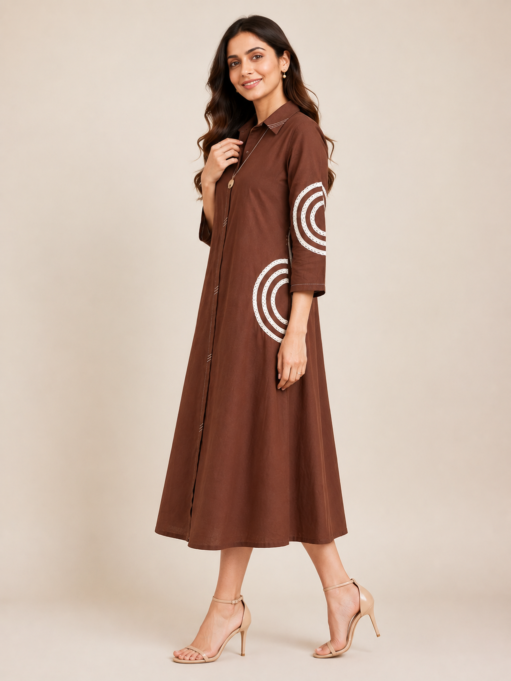 A woman modeling a brown midi shirt dress with white concentric circle patterns on the sleeves, paired with beige high-heeled sandals against a solid beige studio background.